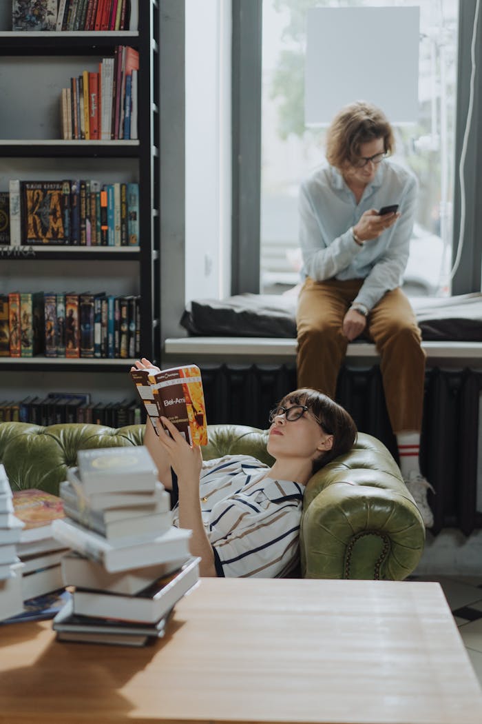 Two young adults in a cozy library setting, one reading and another browsing on a phone by the window.