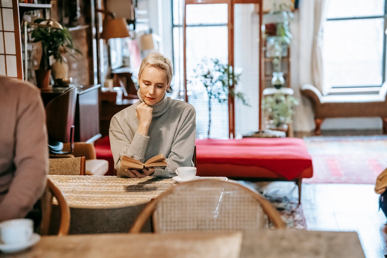 Pensive young adult lady with short blond hair in warm sweater reading interesting book sitting at table during weekend at home with crop unrecognizable husband drinking coffee