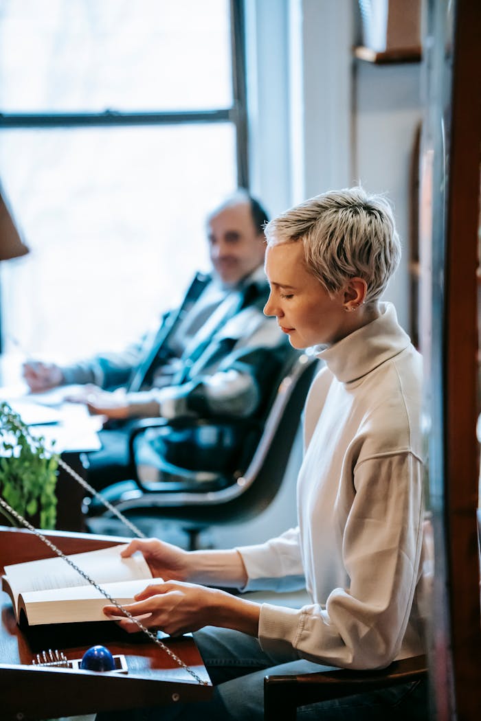 Side view of focused female with short blond hair sitting at table and reading book against blurred man in light room in daytime